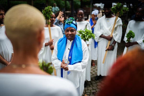 A woman in a blue headscarf with her hands clasped stands in front of other women in white robes and carrying signs. 